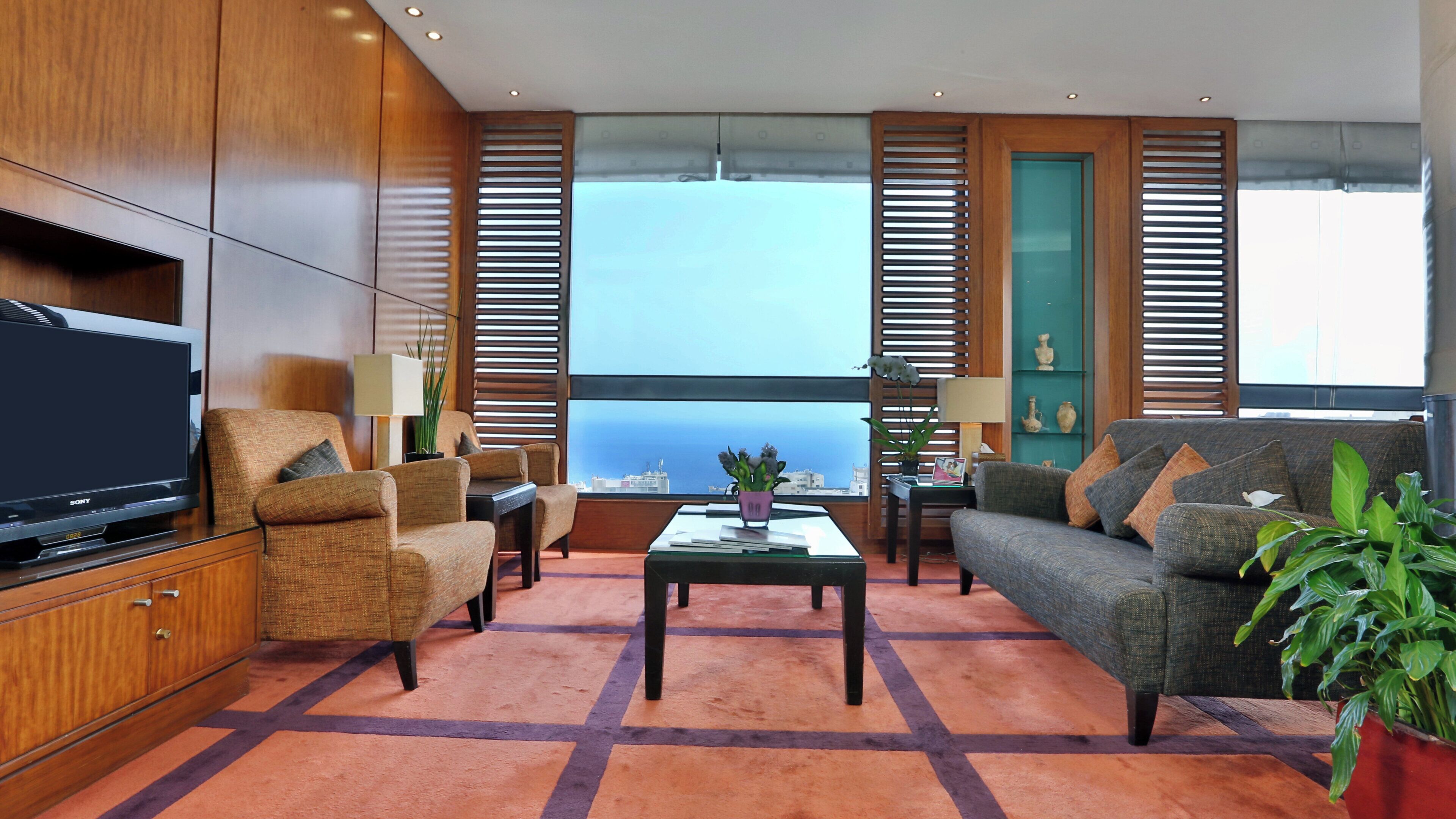 A modern living room with a flat-screen TV, comfortable seating, a coffee table, and large windows with blinds.