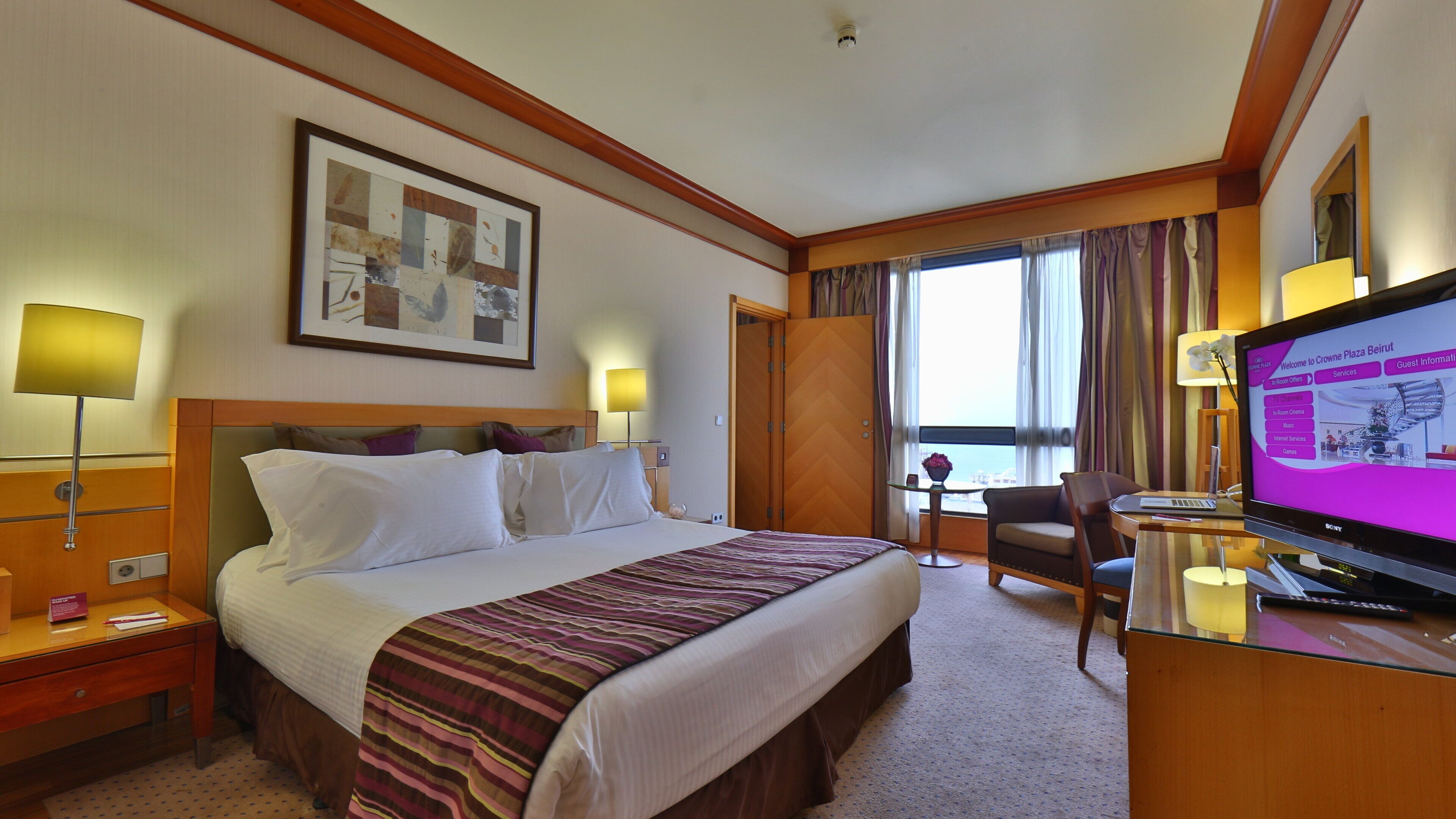 A hotel room with a bed, a desk with a computer, a TV, and a window with curtains.