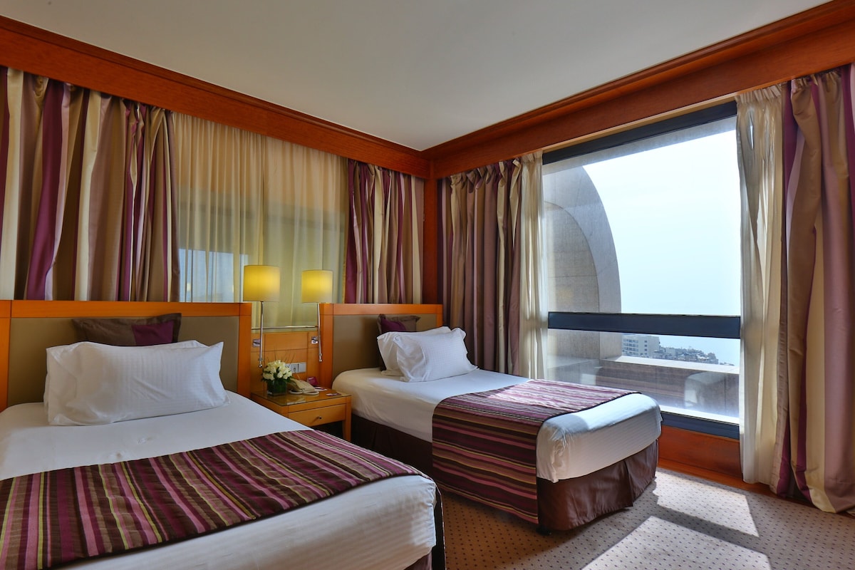 A hotel room with two beds, a nightstand, a lamp, and a window with curtains.