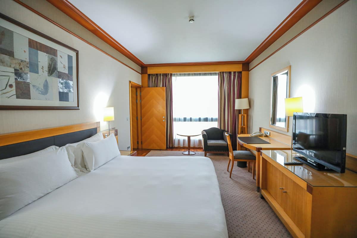 Quarto de hotel com uma cama grande, uma escrivaninha com televisão, uma cadeira e uma janela com cortinas.