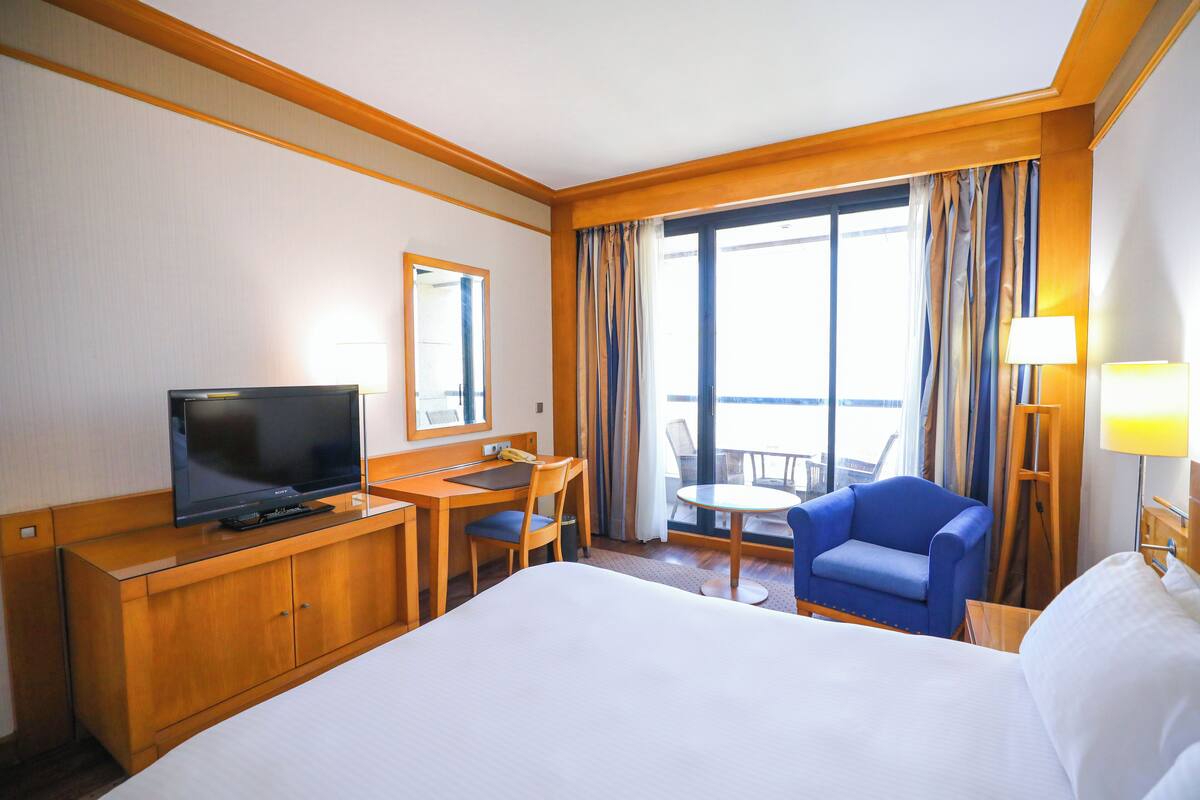 Quarto de hotel com uma cama grande, uma escrivaninha com televisão, uma cadeira, uma mesinha e uma varanda com cortinas.
