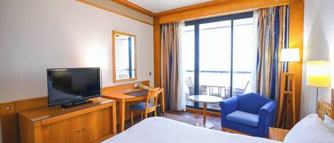 A hotel room with a large bed, a desk with a television, a chair, a small table, and a balcony with curtains.