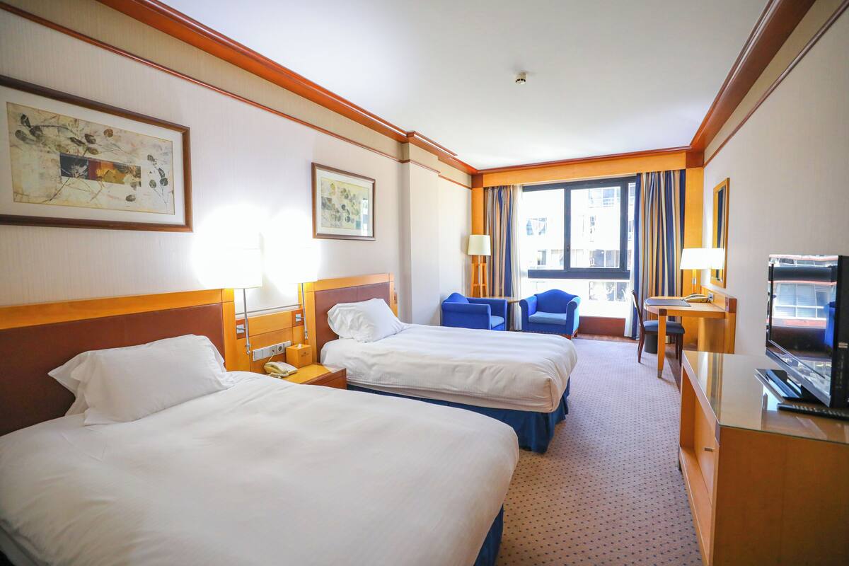 Quarto de hotel com duas camas, uma escrivaninha, uma televisão e uma janela com cortinas.