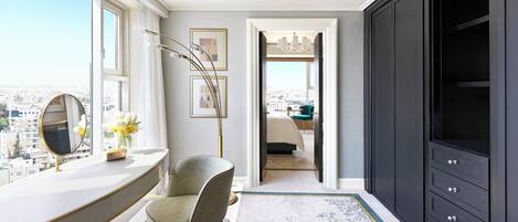 A modern bedroom with a city view, a large wardrobe, and a vanity with a mirror and flowers.