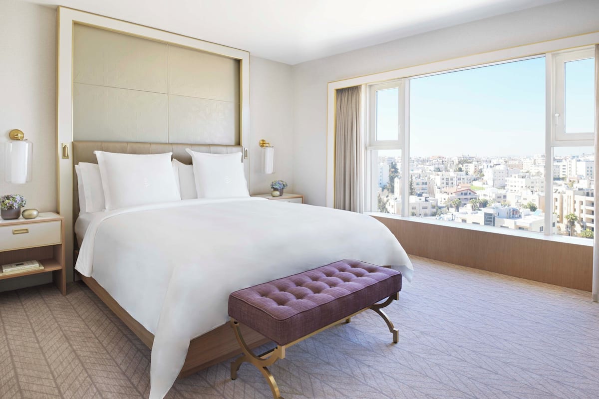 A modern hotel room with a large bed, a bench, a nightstand, and a view of the city through a large window.