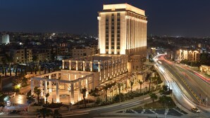 A hotel with a prominent tower, a landscaped area with palm trees, and a busy road with light trails.
