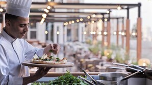 A chef holding a pizza on a wooden board, with a kitchen and dining area in the background.