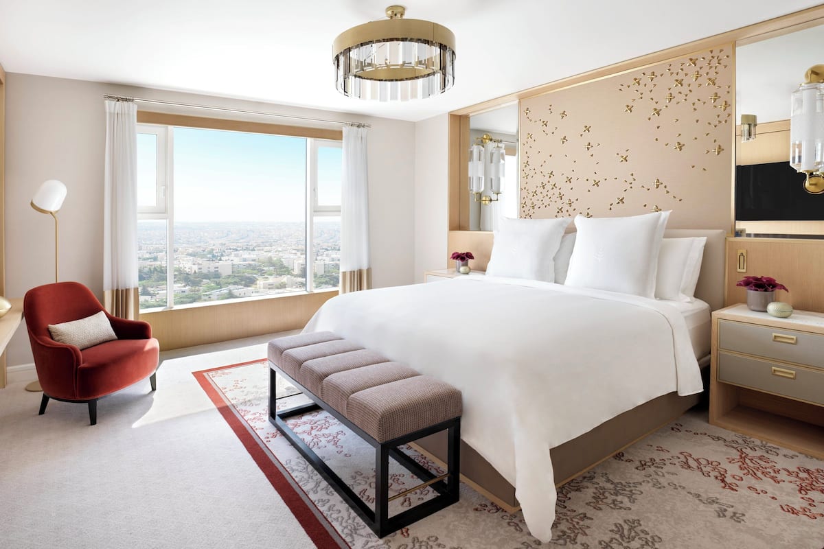 A modern hotel room with a large bed, a red armchair, a bench, a large window with a city view, and a decorative headboard.