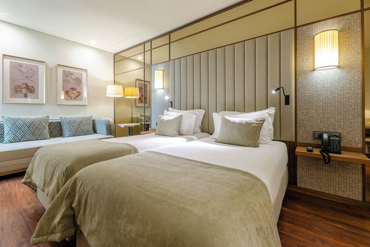 A hotel room with two beds, a sofa, a bedside table with a lamp, and artwork on the walls.