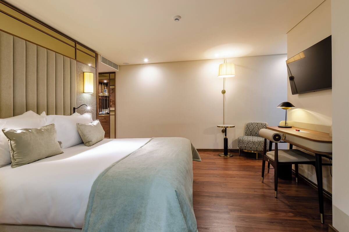 A hotel room with a bed, a desk, a chair, a TV, and a lamp.