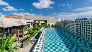 Una piscina en la azotea con diseño a cuadros, rodeada de sillones de relax y sombrillas, bajo un cielo azul despejado.
