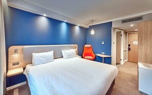 A hotel room with a bed, a bedside table, a chair, a small table, and a door.