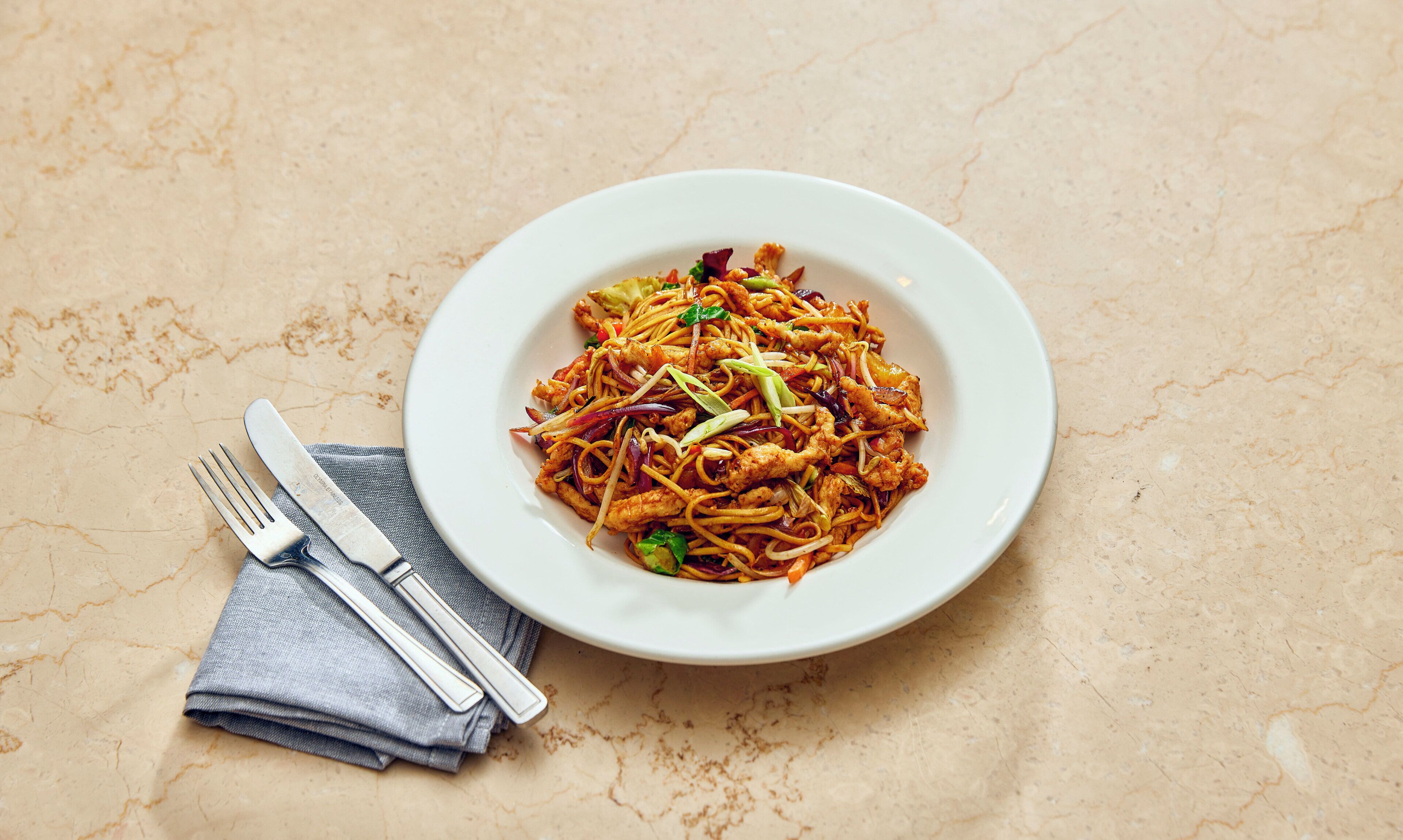 A plate of stir-fried noodles with vegetables and meat, a fork, and a napkin on a textured surface.