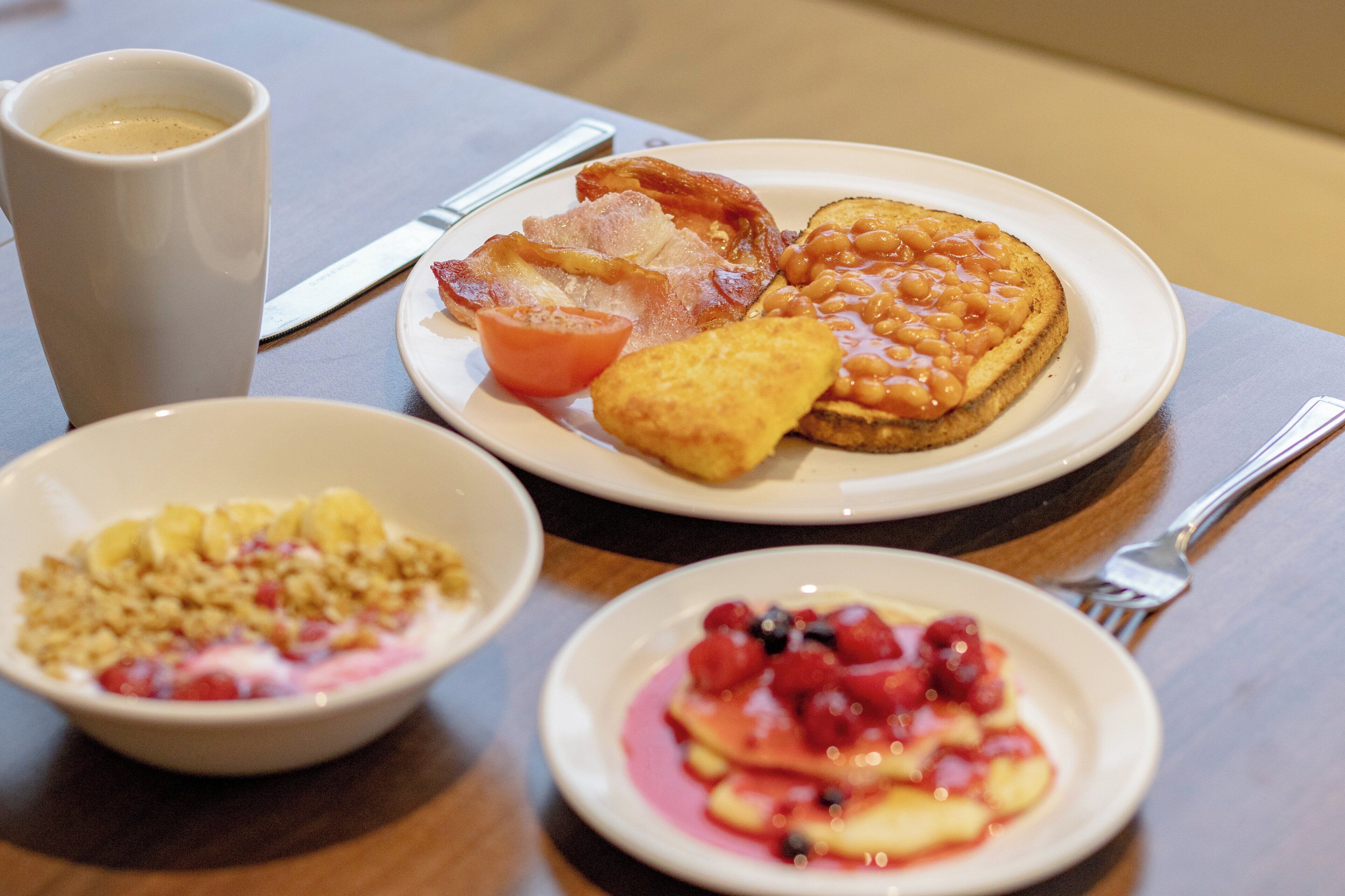A breakfast setting with a plate of toast, beans, bacon, and a tomato, a bowl of cereal, and a plate of pancakes with berries.