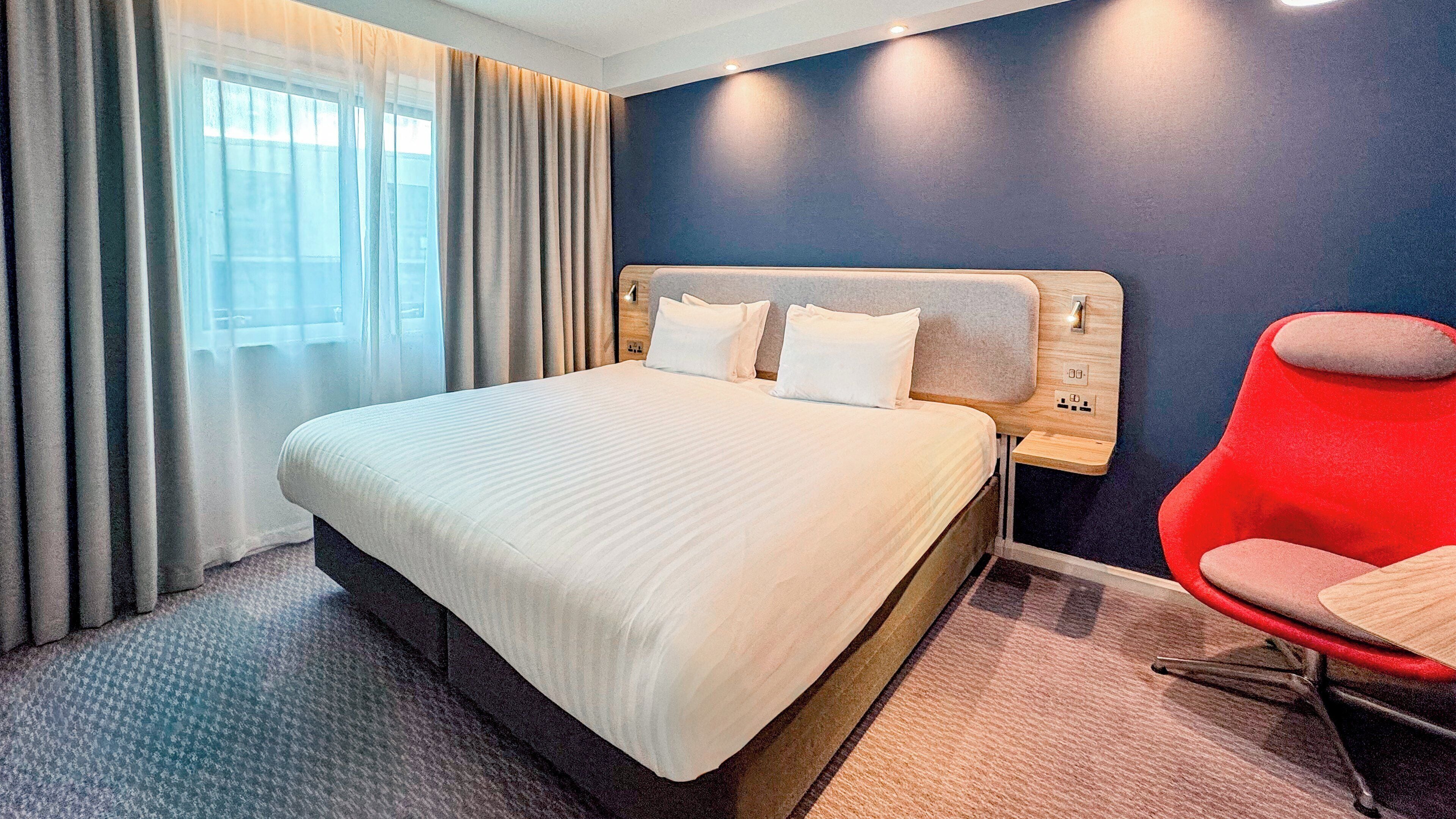 A hotel room with a large bed, a red chair, and a view of the outdoors through the window.