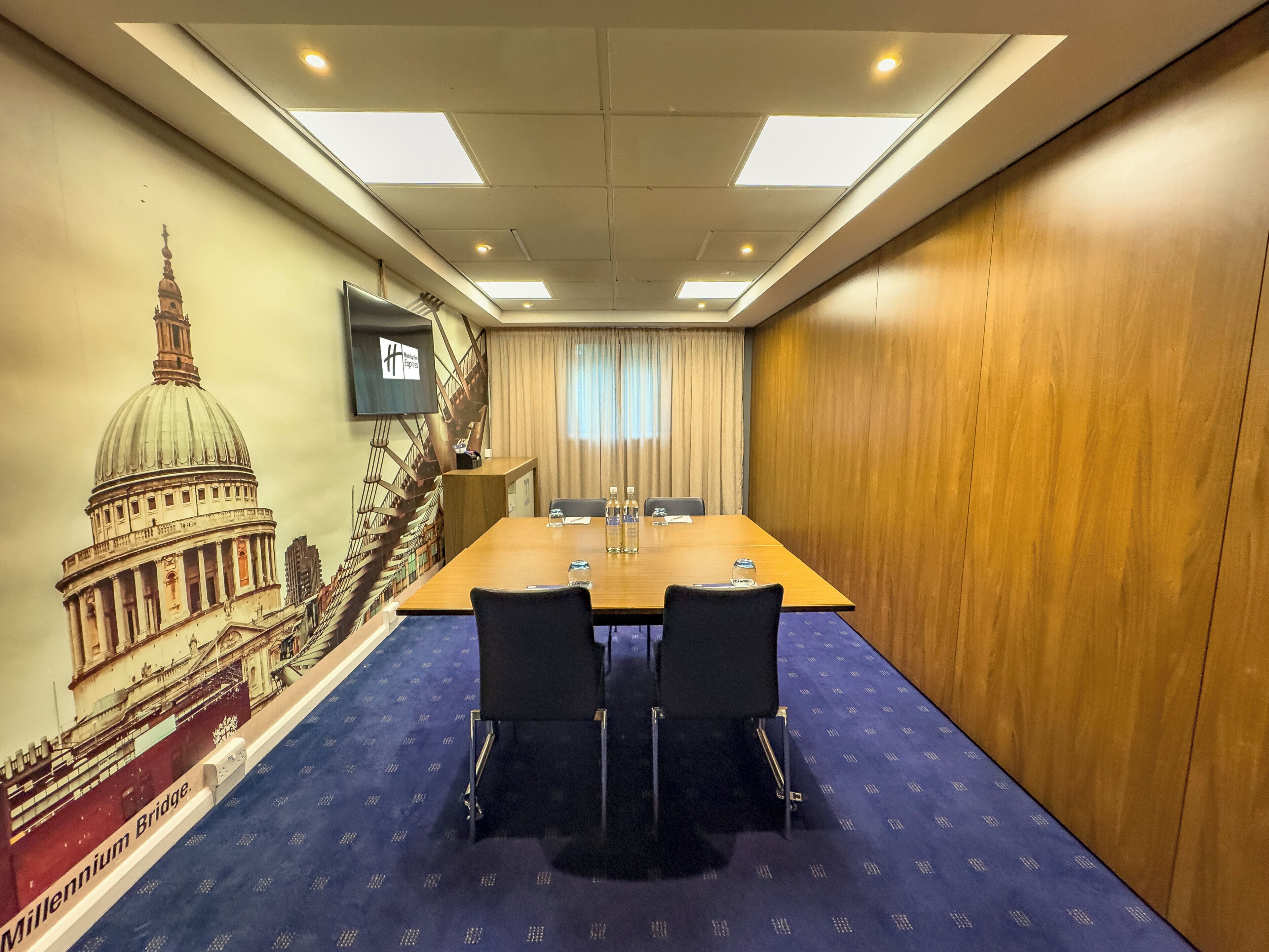 Uma sala de conferências com uma mesa comprida, duas cadeiras, uma tela de televisão e um mural de um edifício histórico.