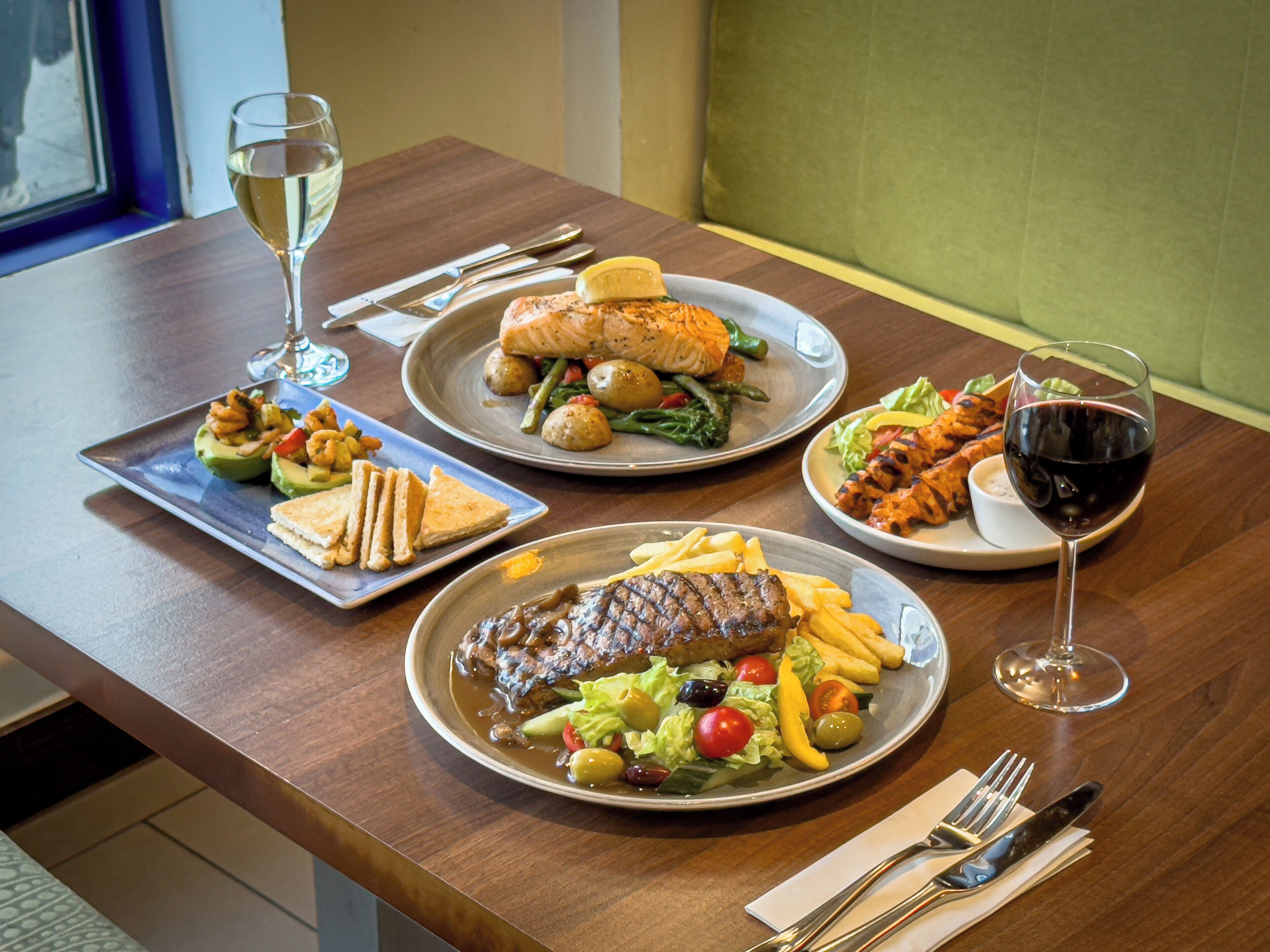 A dining table with three plates of food, two glasses of wine, and cutlery.