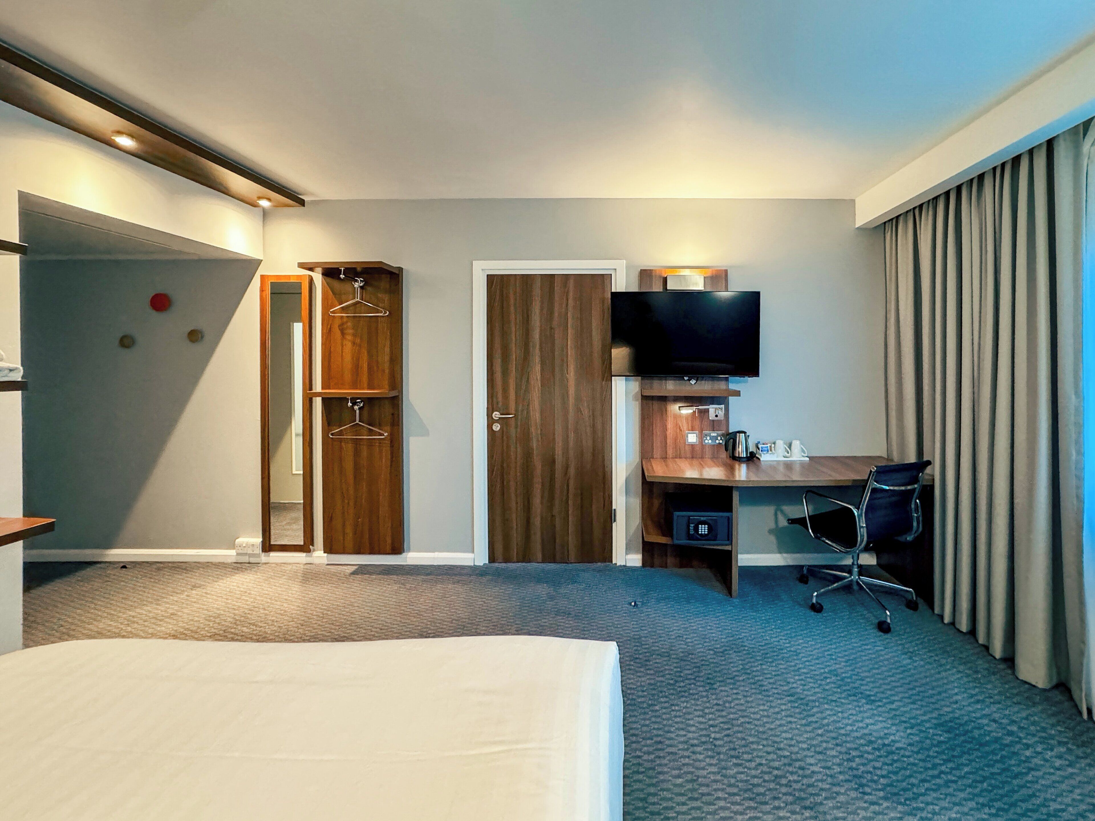 A hotel room with a bed, a desk with a chair, a TV, and a wardrobe with hangers.