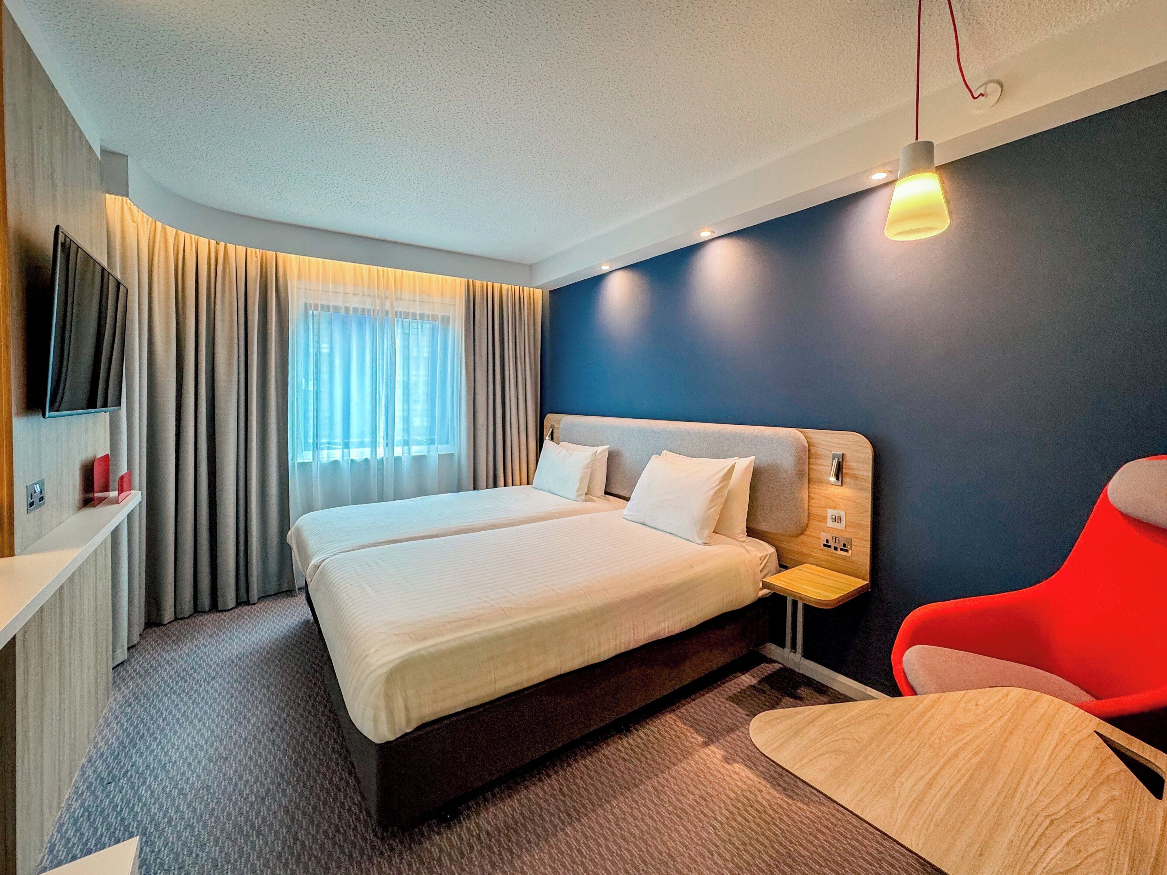 A modern hotel room with a large bed, a TV, a desk, and a red chair.
