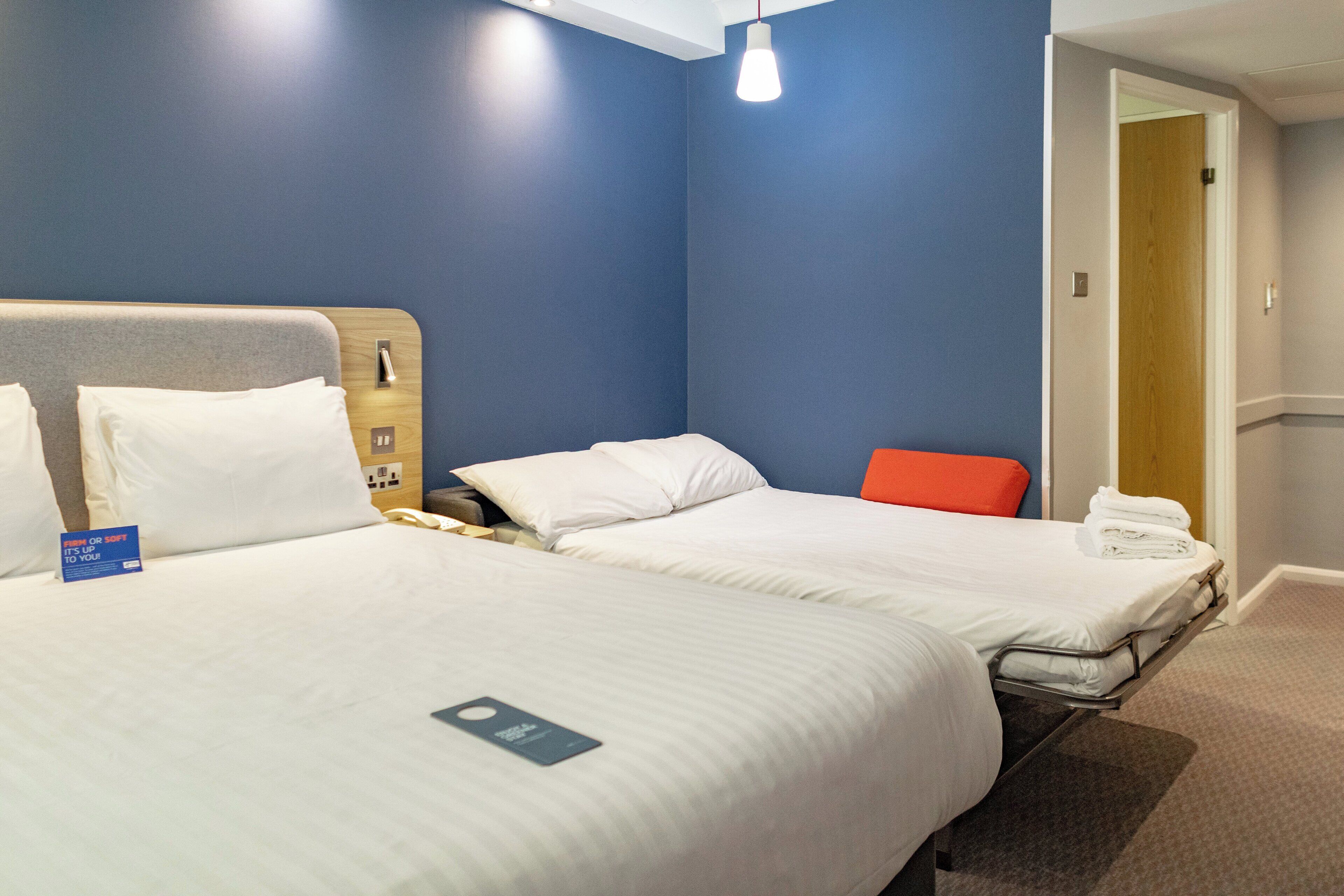 Quarto de hotel com duas camas, uma cabeceira de madeira, uma cadeira vermelha e uma porta que dá para outro quarto.