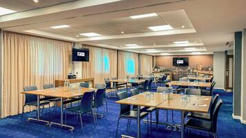 A hotel conference room with multiple tables and chairs, a flat-screen TV, and a cityscape mural on the wall.