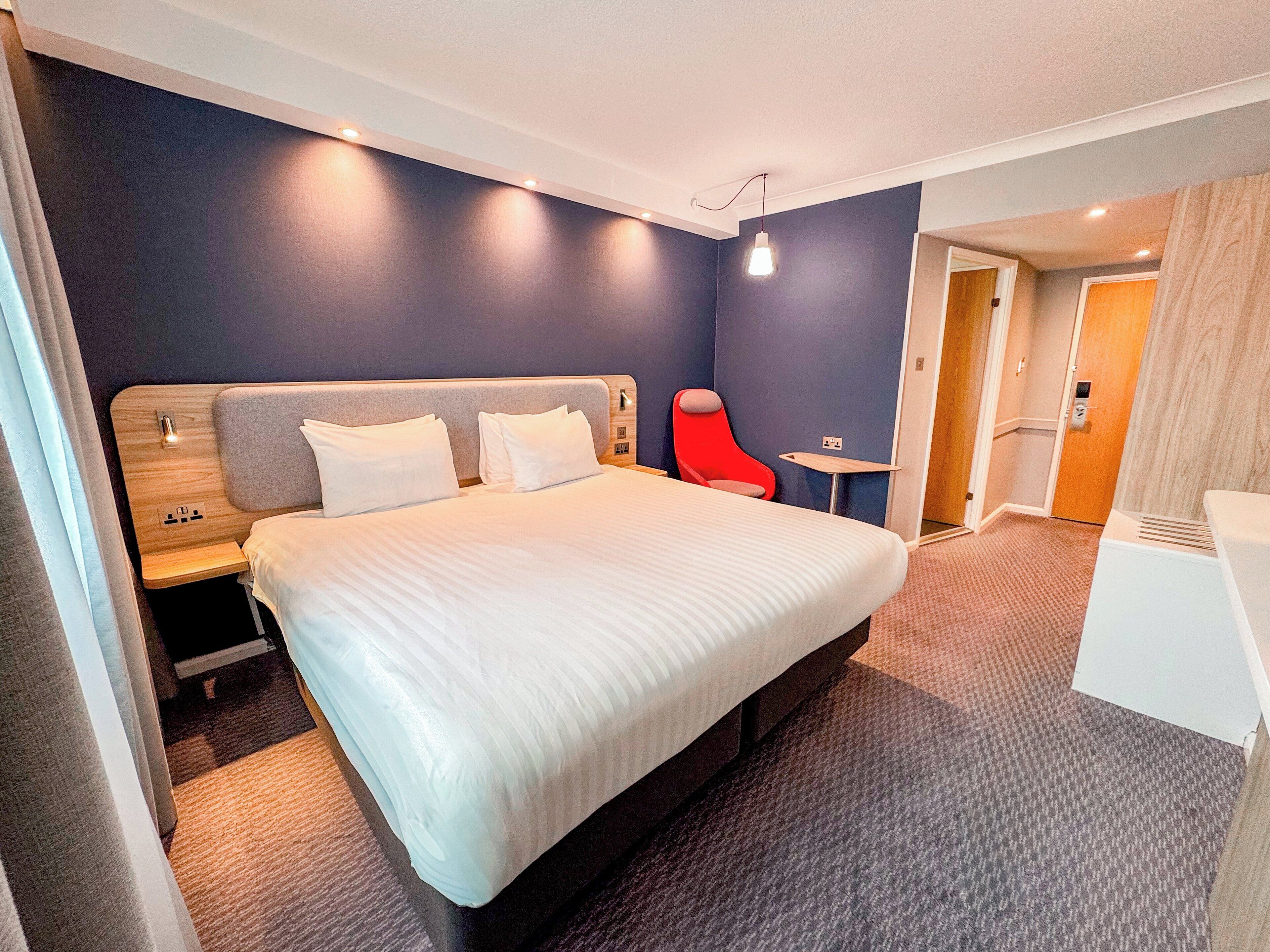 A modern hotel room with a large bed, a small table, a red chair, and a separate desk area.