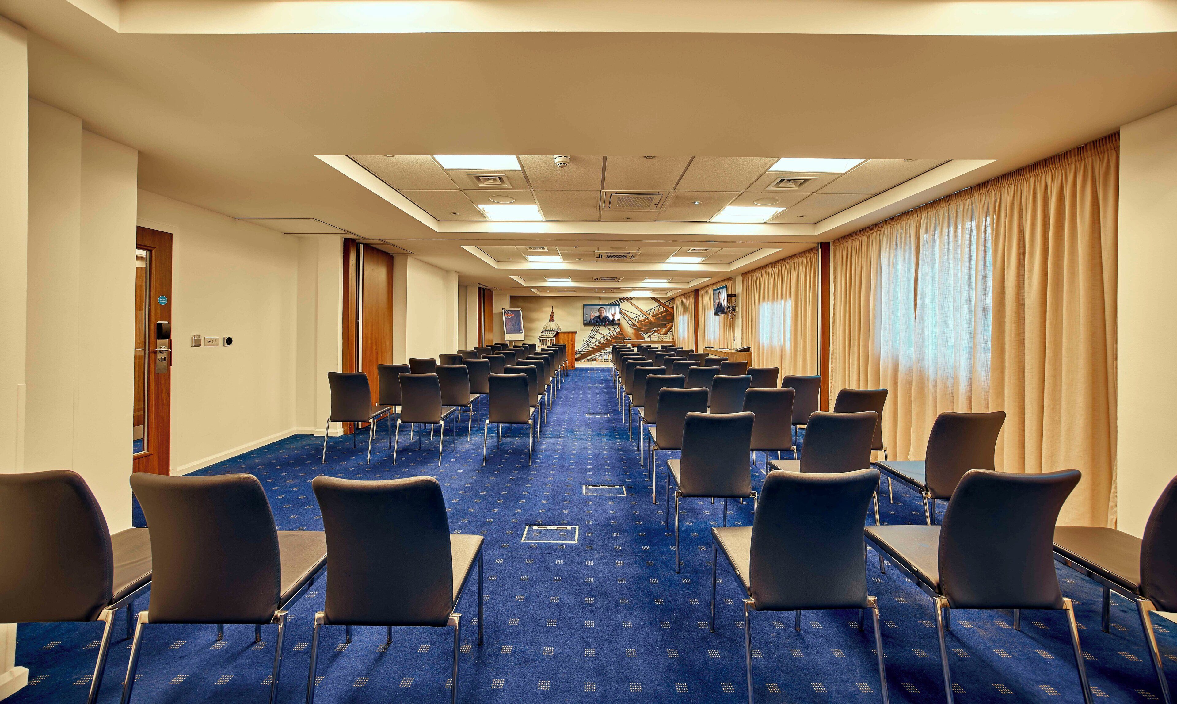 A hotel conference room with rows of chairs, a blue carpet, and a large window with curtains.