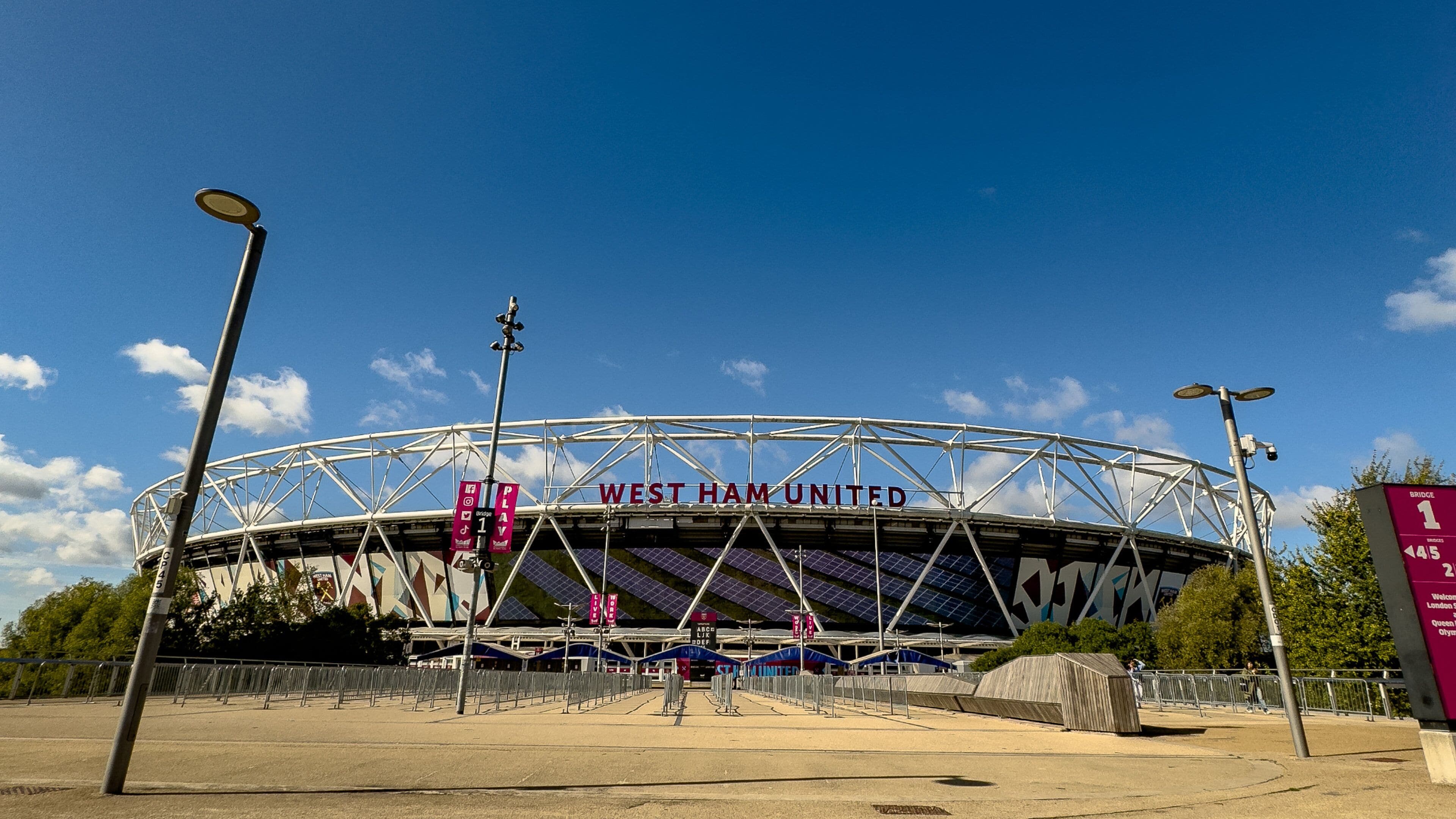 A stadium with 'West Ham United' written on the facade, surrounded by a fence, under a clear sky.