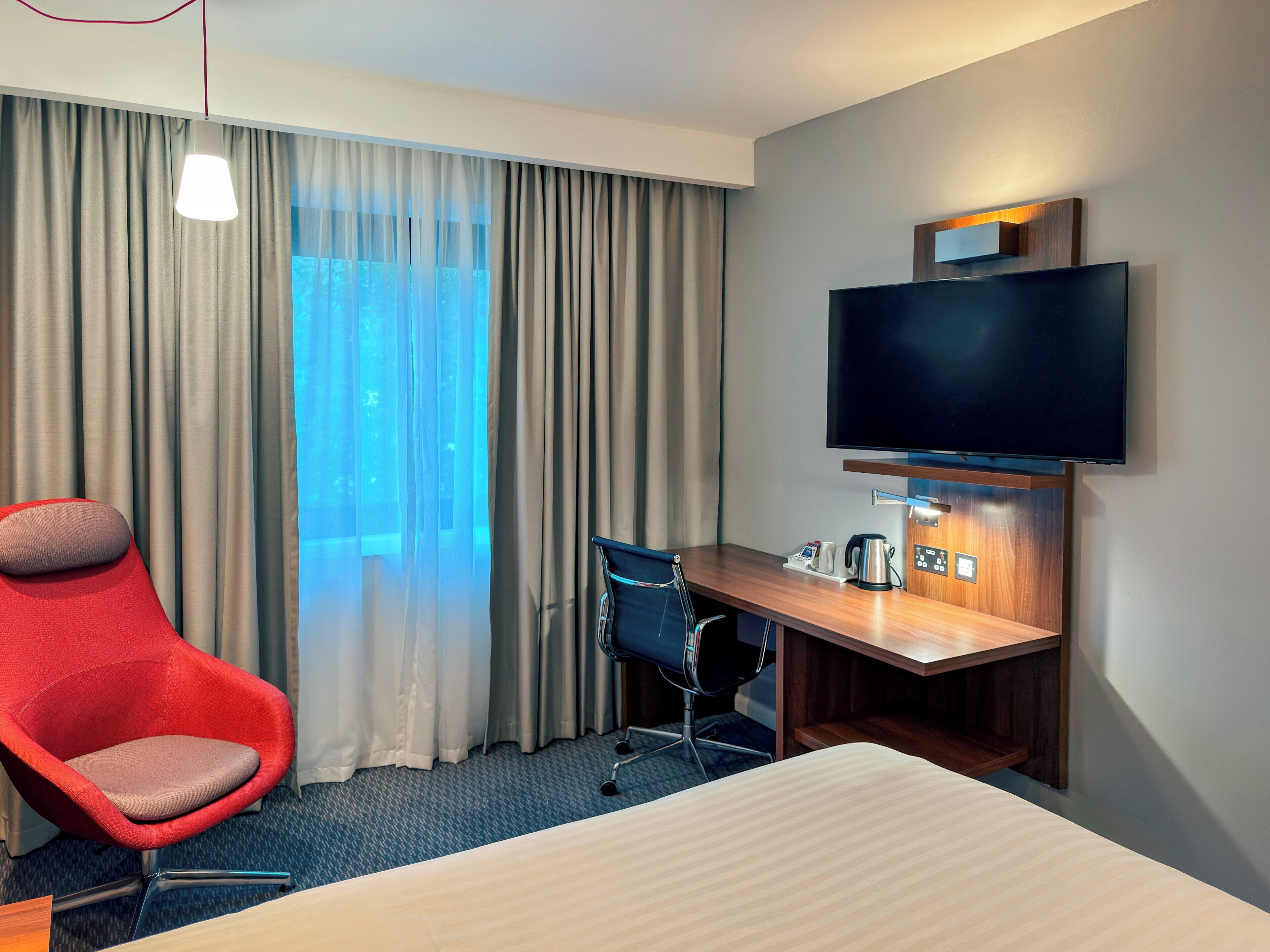 Ett hotellrum med en säng, ett skrivbord med en platt-TV, en röd stol, en blå kontorsstol och ett stort fönster med gardiner.