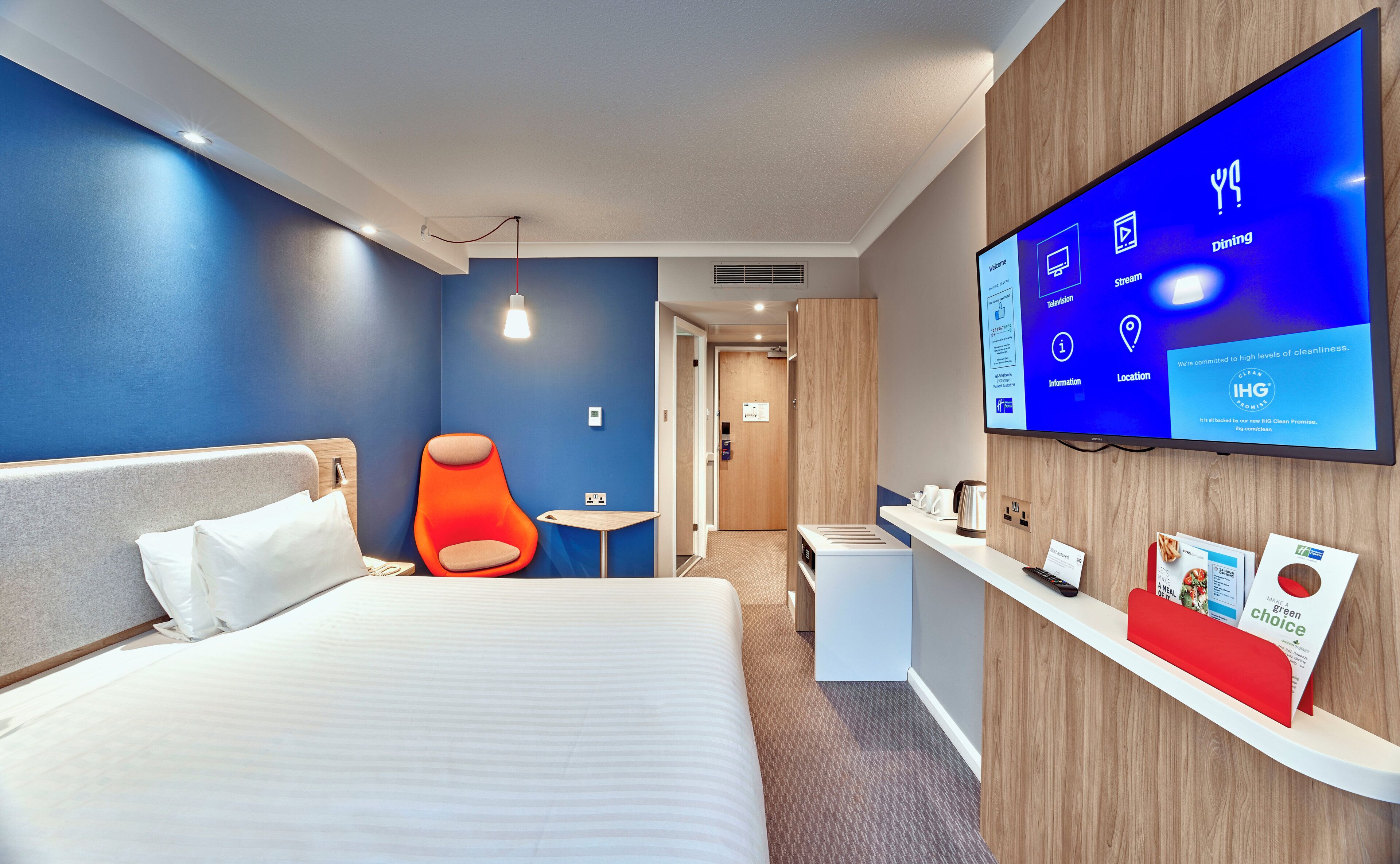 Una habitación de hotel moderna con una cama, un sillón naranja brillante, un televisor mostrando información de la habitación y un mostrador de recepción con un menú.
