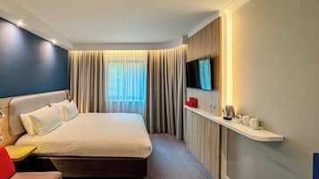 A hotel room with a bed, a TV mounted on the wall, a small table, and a shelf with items.