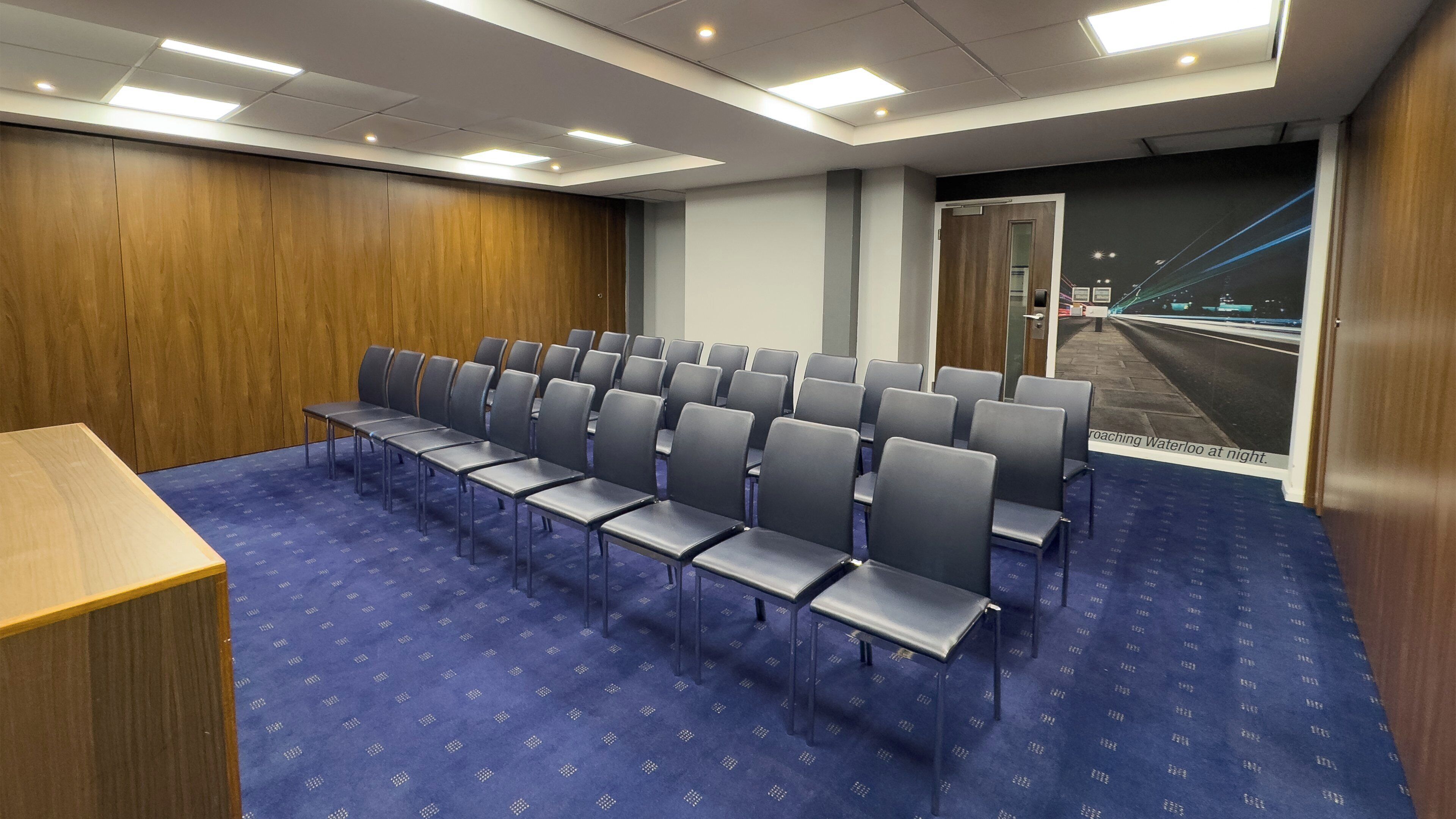 A conference room with rows of chairs and a podium, a blue carpet, wooden panel walls, and a night view poster.
