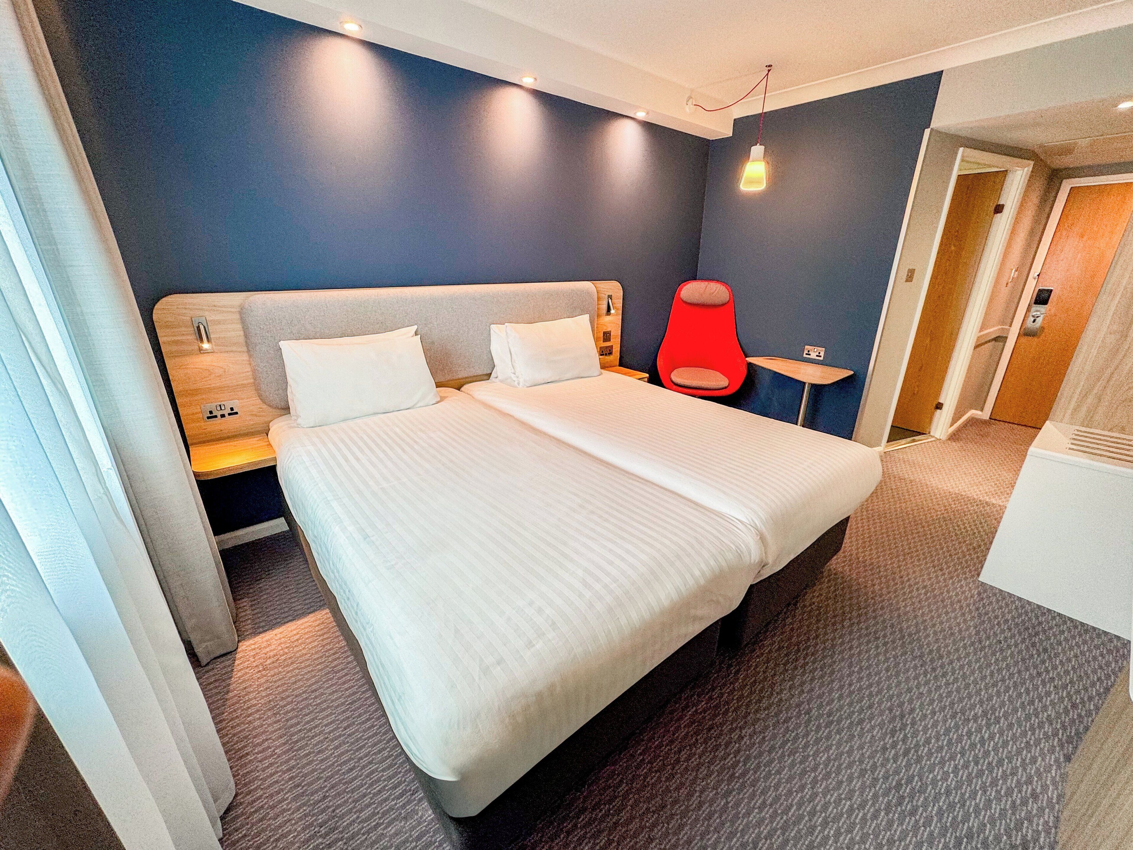 A modern hotel room with a large bed, a bedside table, a red chair, and a small table.