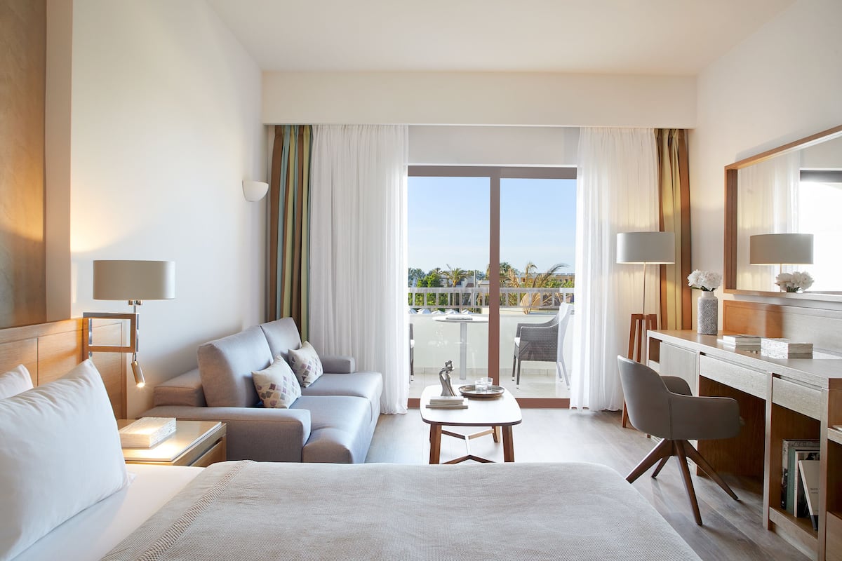 A hotel room with a bed, a sofa, a desk, and a chair. There is a balcony with a view of the outside.