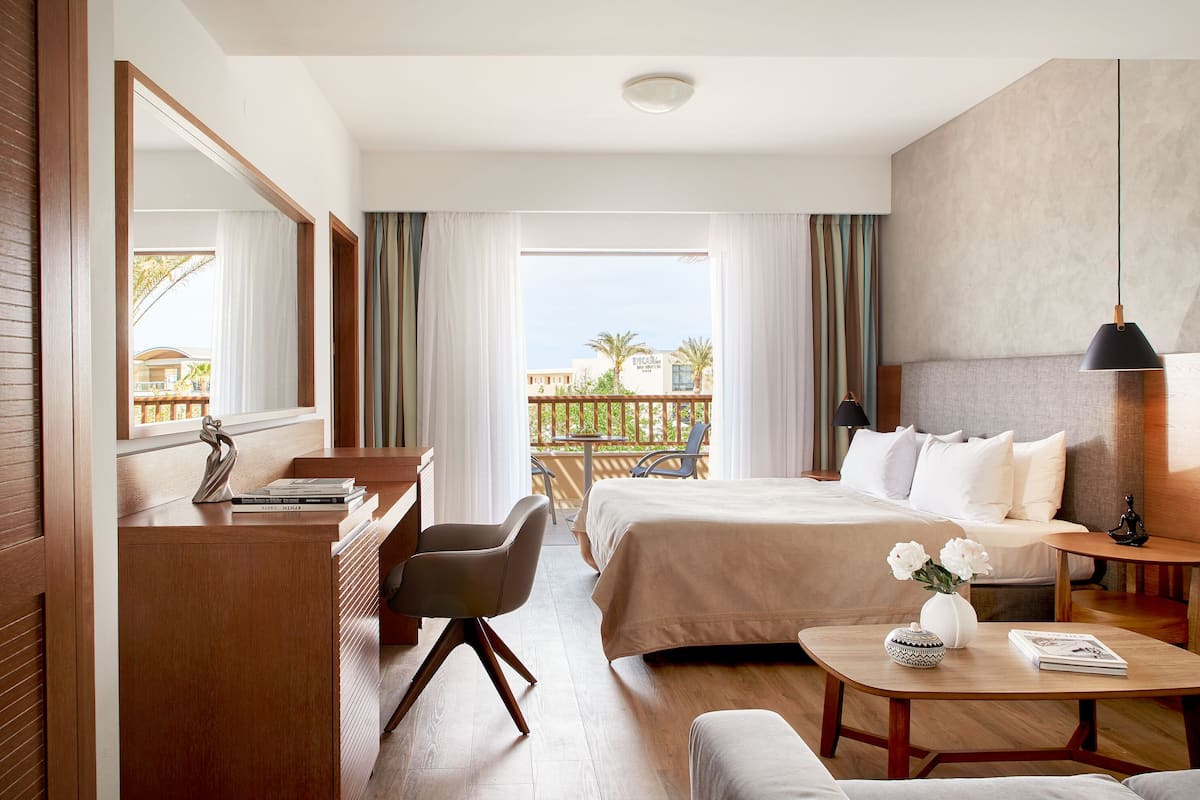 A hotel room with a bed, a desk, a chair, a small table, and a balcony with a view of palm trees and buildings.