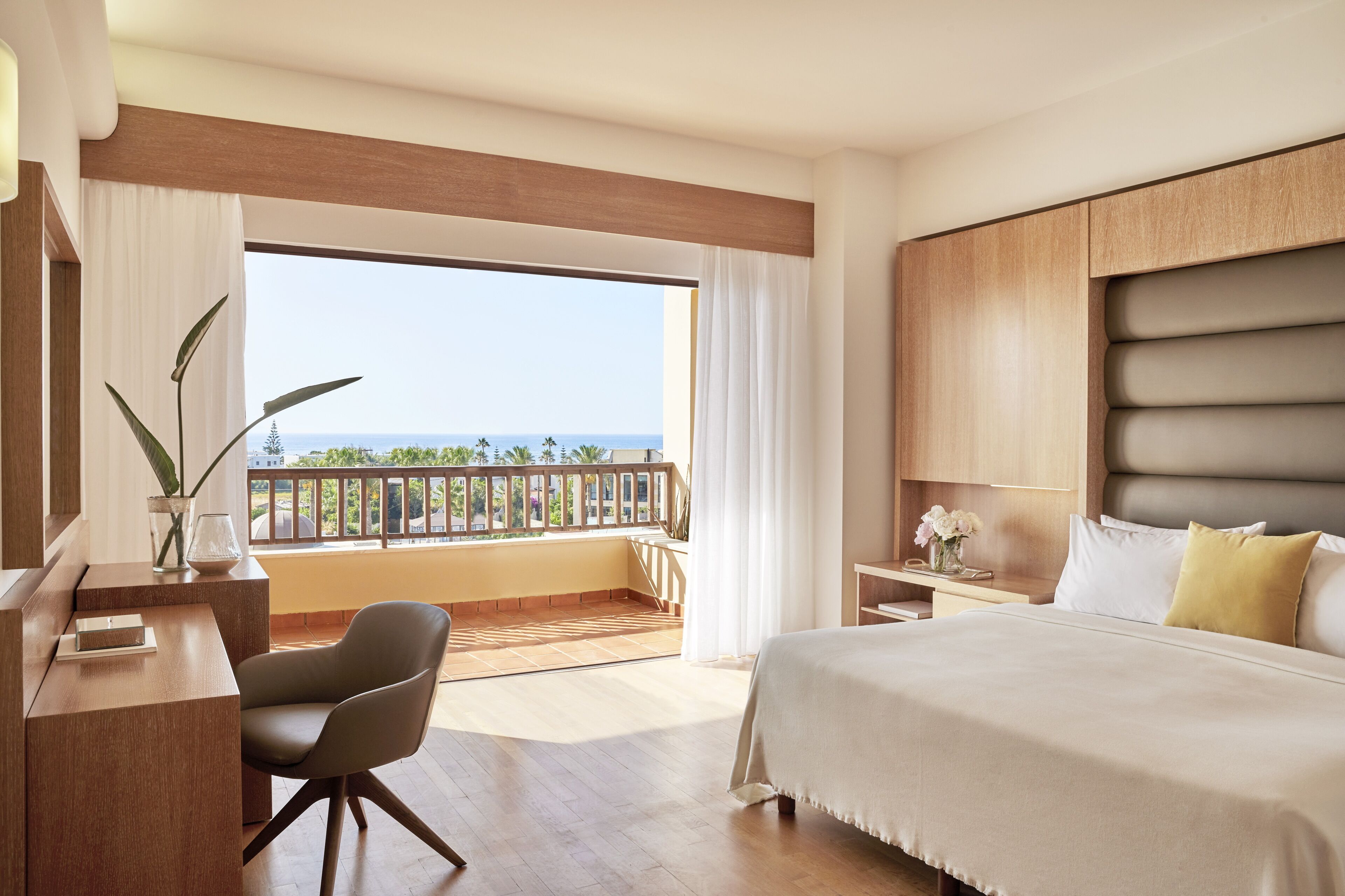 A hotel room with a large bed, a desk, a chair, and a balcony with a view of the ocean.