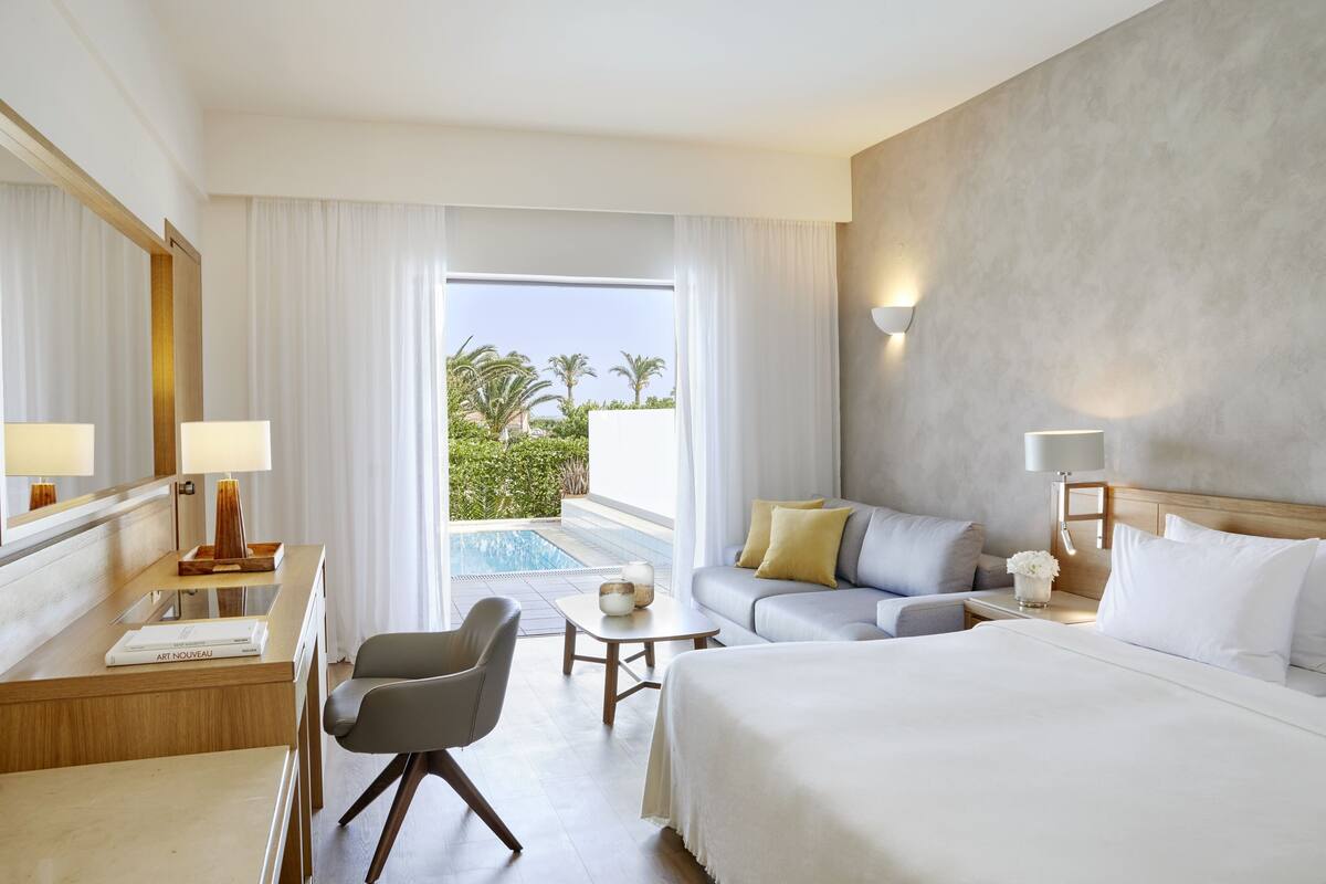 A hotel room with a bed, a sofa, a desk, and a chair. There is a small table and a lamp. The room has a view of a pool and palm trees.