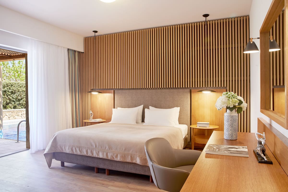 A modern hotel room with a large bed, a wooden headboard, two bedside tables with lamps, a small table with a vase of flowers, and a view of the outdoor pool area.