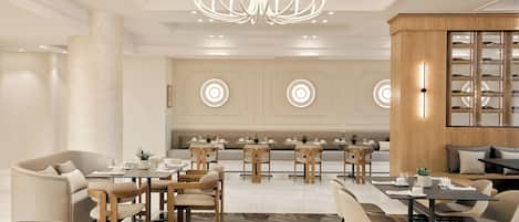 Un vestíbulo de hotel moderno con un área de estar central, varias mesas de comedor y un candelabro.
