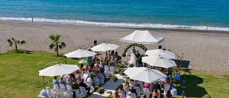 Une cérémonie de mariage sur la plage, avec des invités assis sous des parasols blancs, une allée blanche et une vue dégagée sur l’océan.
