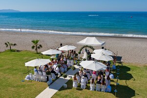A beach wedding ceremony with guests seated under white umbrellas, a white aisle, and a clear view of the ocean.
