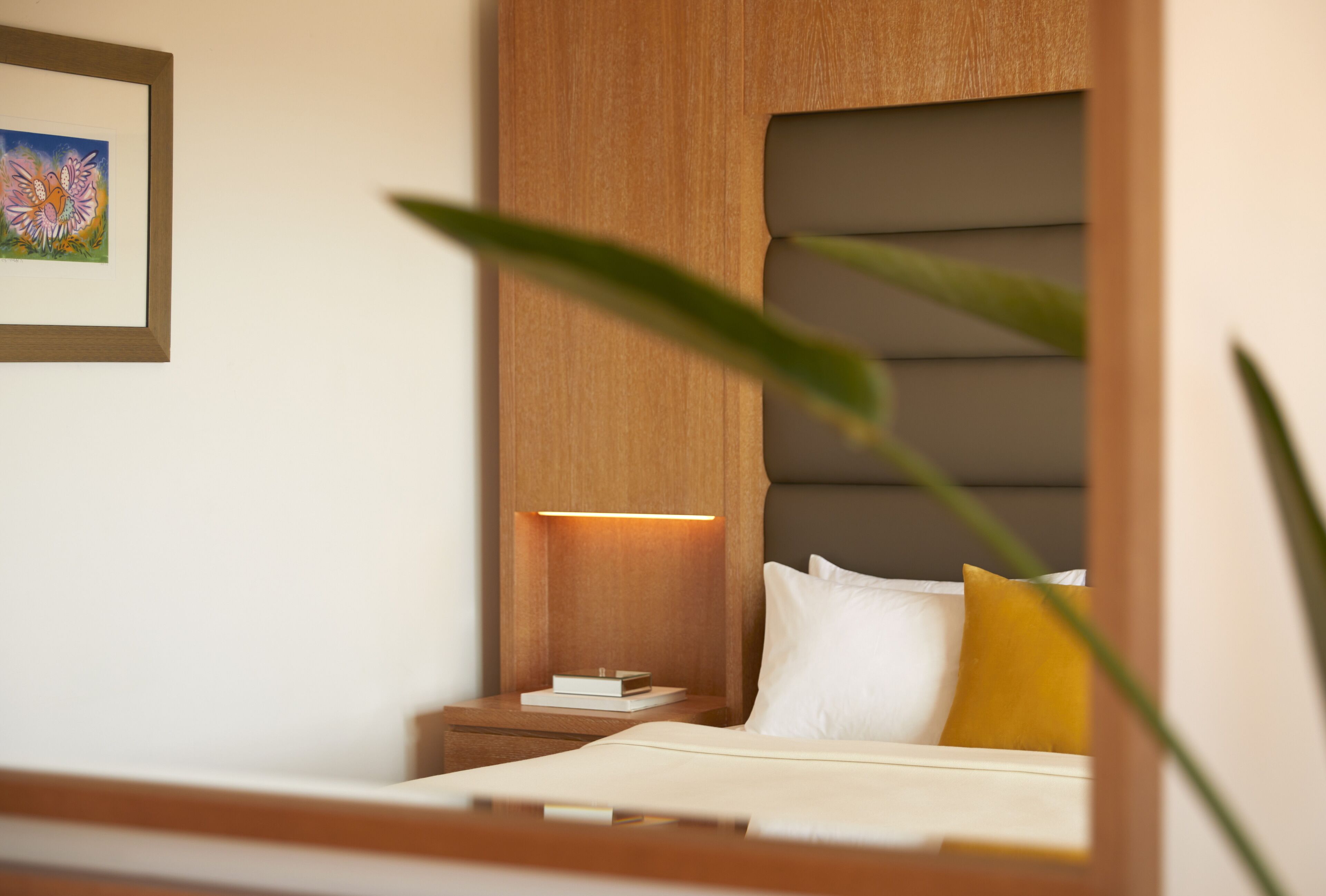 Hotelkamer met een bed, nachtkastje, een tafeltje naast het bed, een ingelijste foto aan de muur en een plant op de voorgrond.
