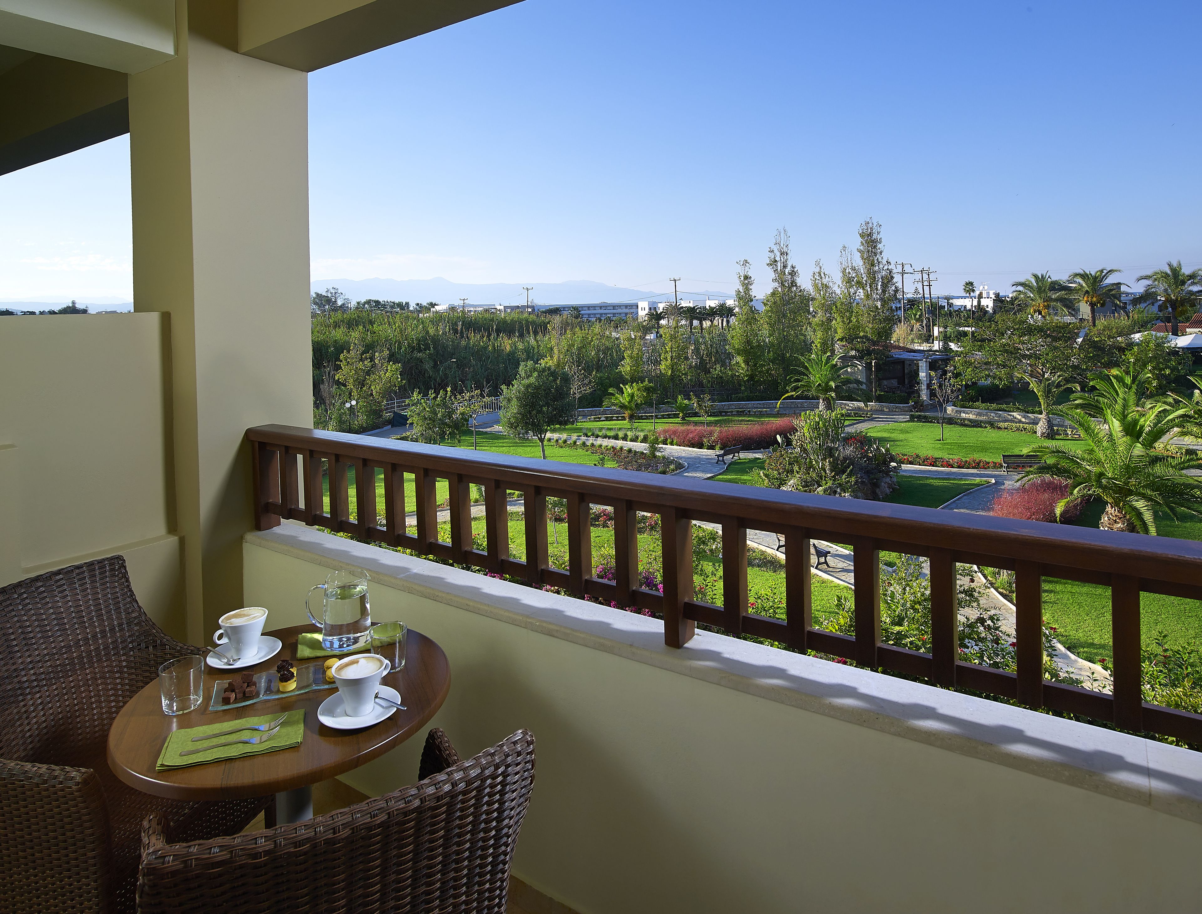 A balcony with wicker furniture, a table with a cup of coffee, and a view of a landscaped garden with trees and a distant mountain range.