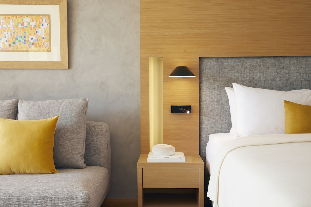 A hotel room with a grey sofa, a bedside table with a lamp, and a bed with white and yellow pillows.