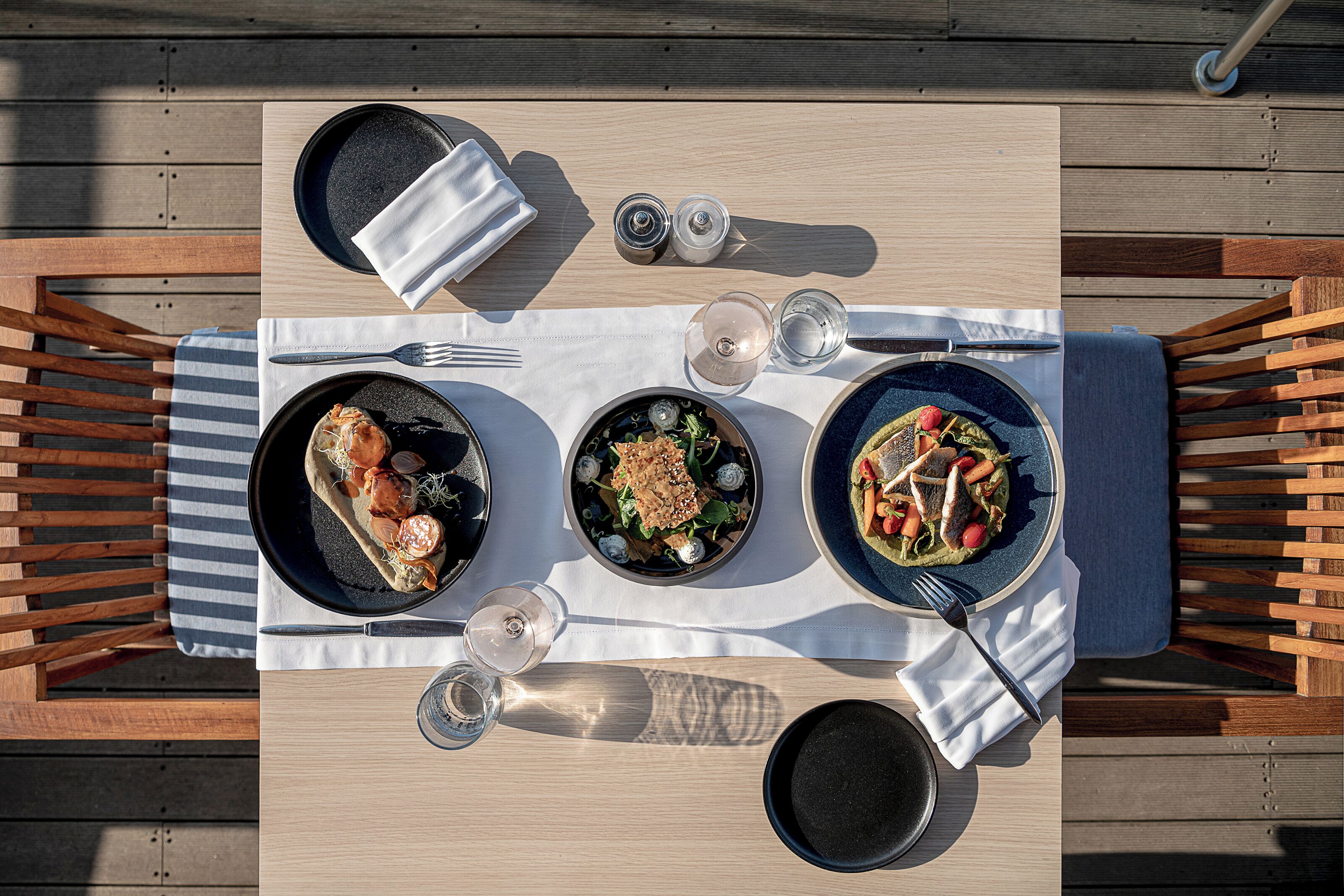 Un tavolo da pranzo con tre piatti di cibo, posate e bicchieri, su un pontile di legno.