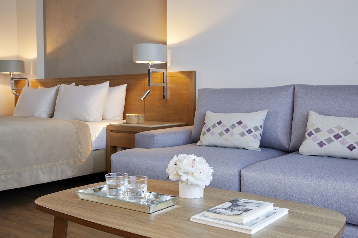 A hotel room with a bed, a sofa, a nightstand with a lamp, a tray with glasses and a vase of flowers on a wooden table.