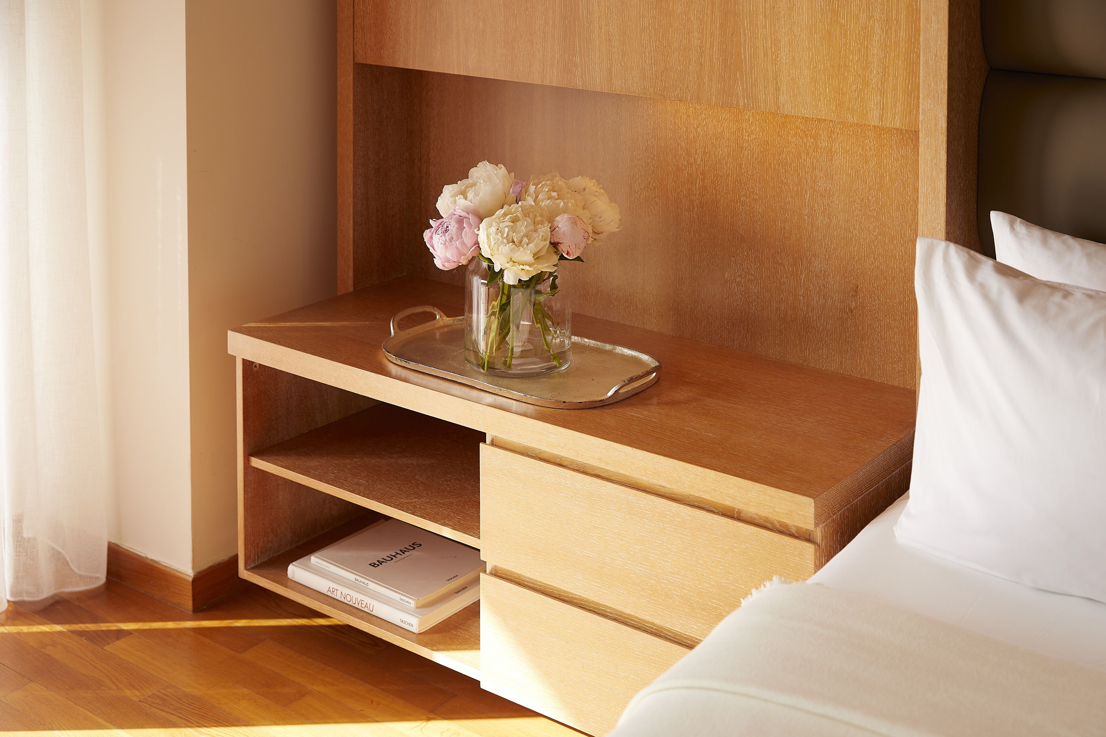 Ein ordentlich eingerichtetes Hotelzimmer mit einem Nachttisch aus Holz, einer Vase mit Blumen, einem Tablett und einem Buch mit dem Titel „ART NOUVEAU“.