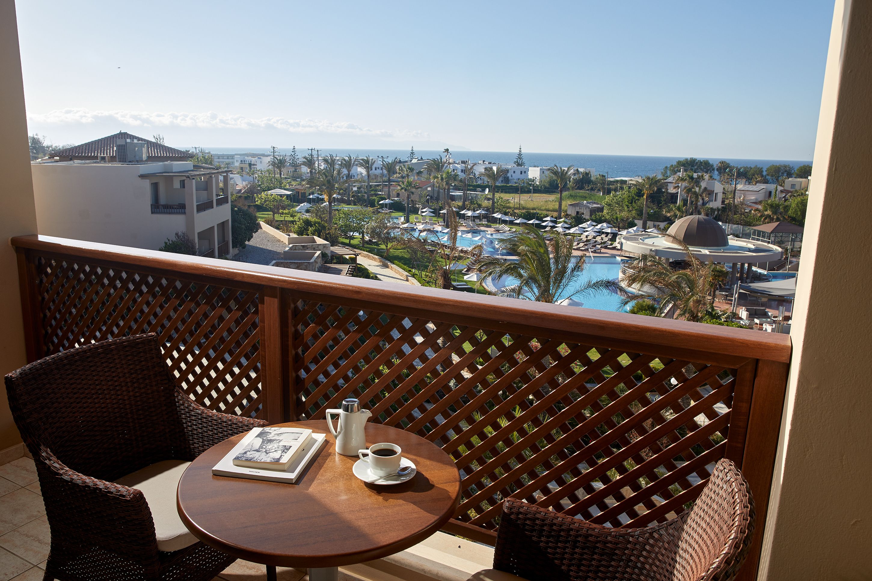 Uma varanda com móveis de vime, uma mesa com cafeteira e xícara, e vista para um resort com piscina e palmeiras.