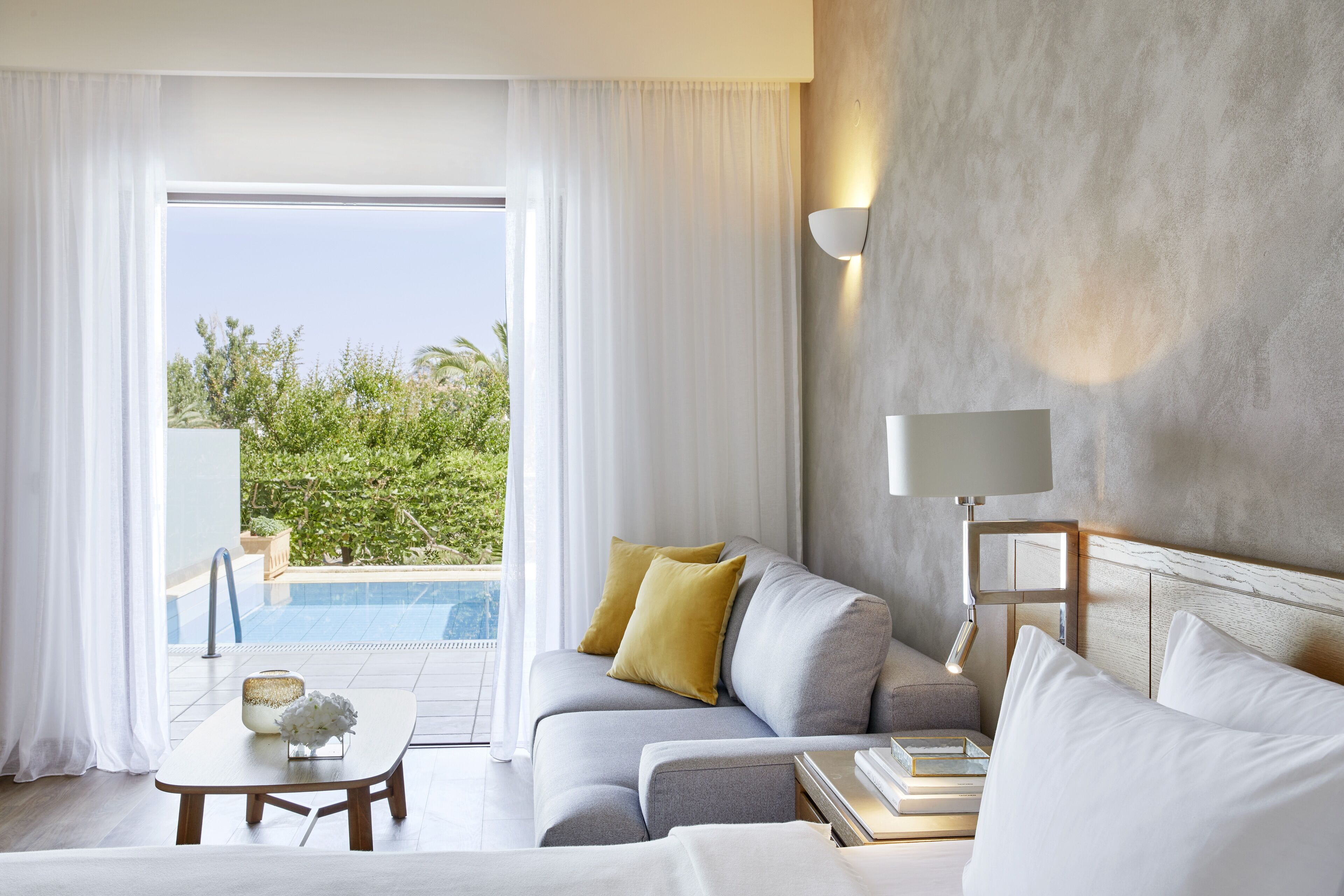 A modern hotel room with a grey sofa, white bedding, a bedside table with books, a lamp, and a view of a pool and greenery.