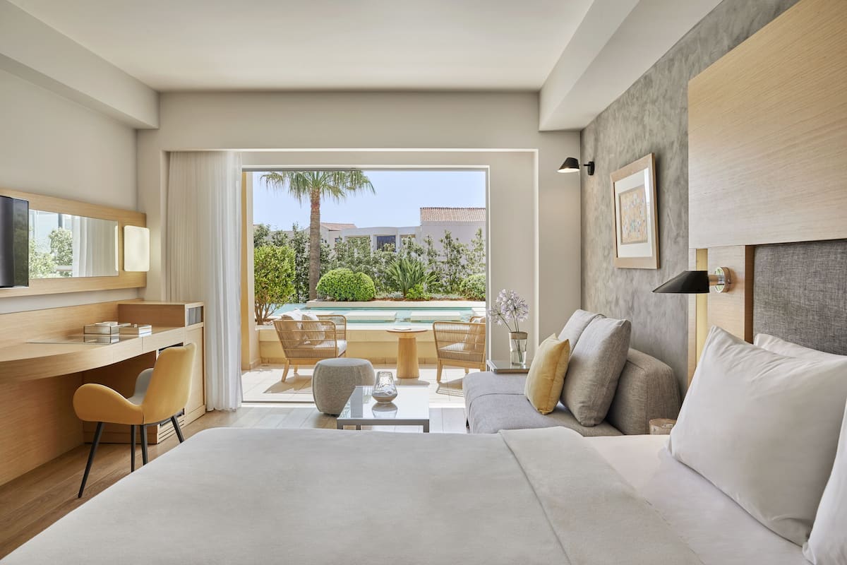 A modern hotel room with a large bed, a desk, and a seating area with a view of a pool and greenery.